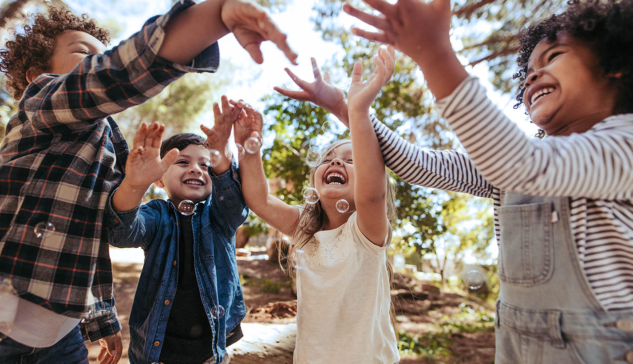 Group of kids playing with soap bubbles in forest. Boy blowing soap bubbles with friends trying to catch the bubbles.